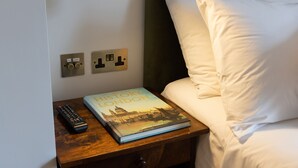 A neatly made bed with white sheets and pillows, a bedside table with a book titled "The History of London" and a remote control, and a wall-mounted light fixture. - THE KNIGHT OF NOTTINGHILL (London)