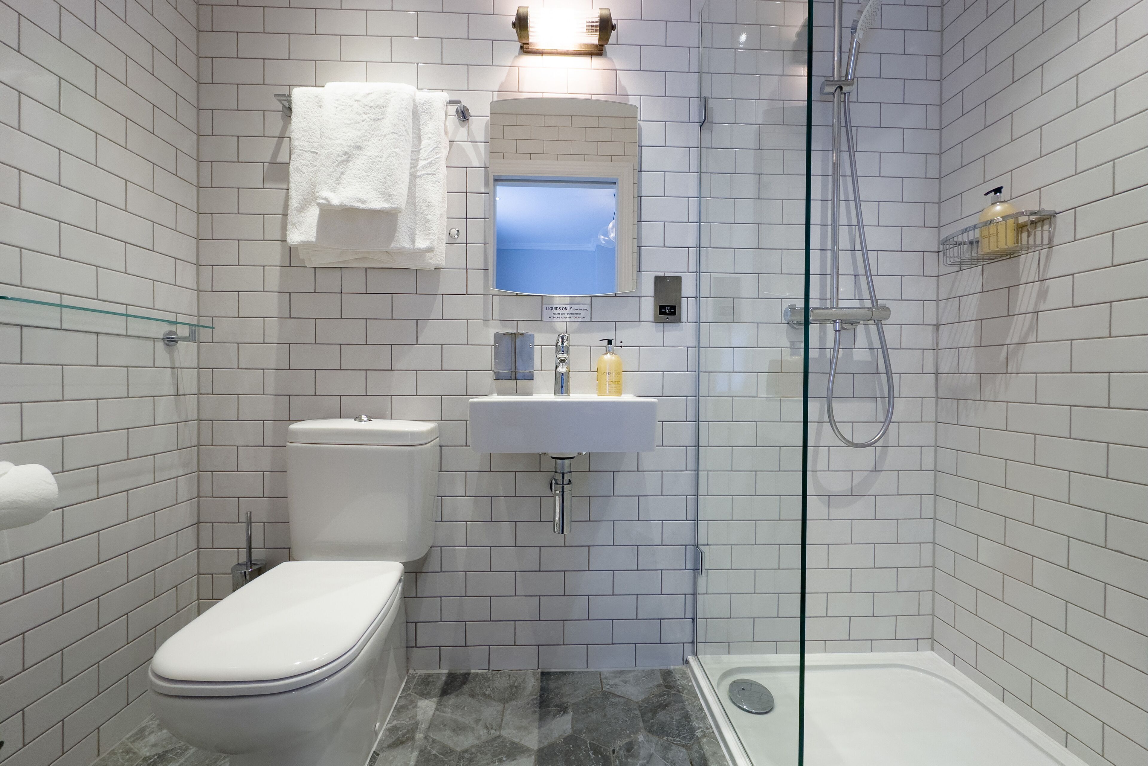 Un bany modern amb rajoles blanques, un vàter, un lavabo amb un mirall a sobre i una zona de dutxa amb envàs de vidre.