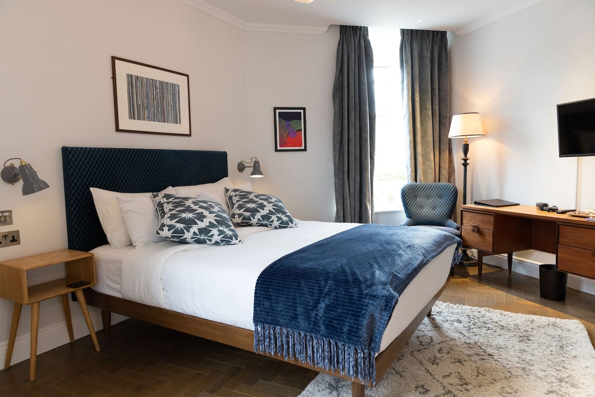 A modern hotel room with a large bed, a desk, a chair, a TV, and artwork on the walls.