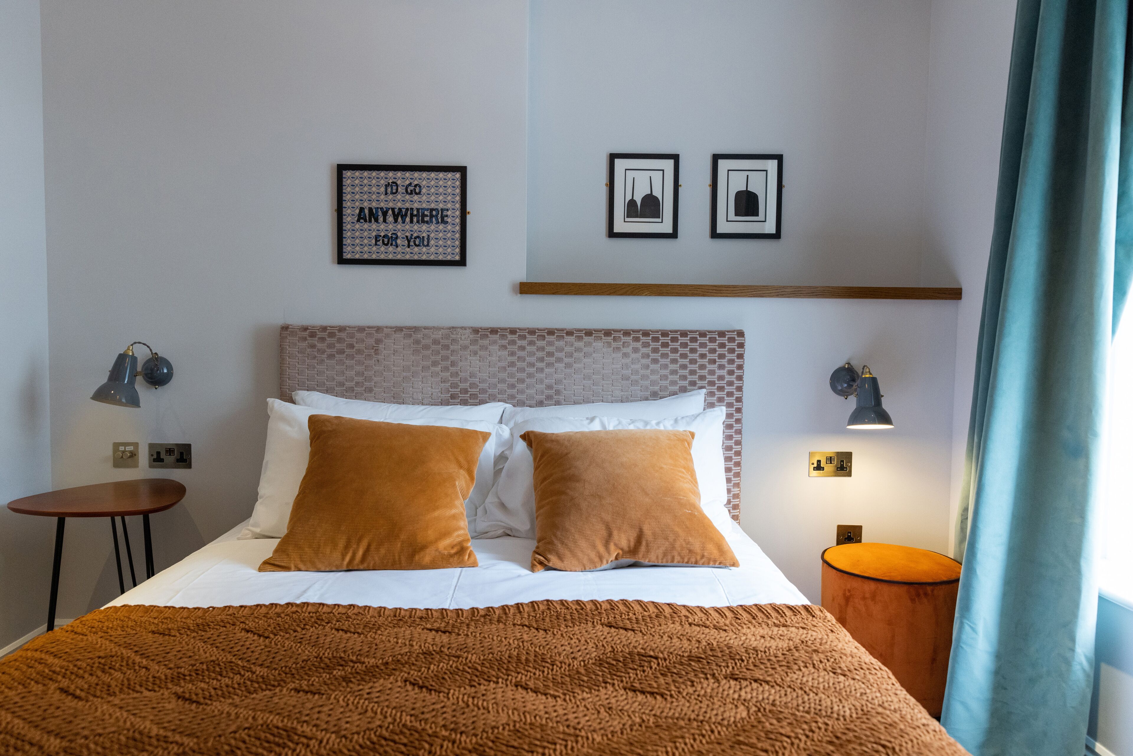 A neatly made bed with two pillows, a bedside table with a lamp, and a small round table with a lamp. There are framed pictures on the wall and a window with curtains.