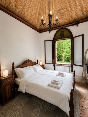 Une chambre à coucher avec un grand lit, un lustre, une fenêtre donnant sur un paysage verdoyant et un miroir.