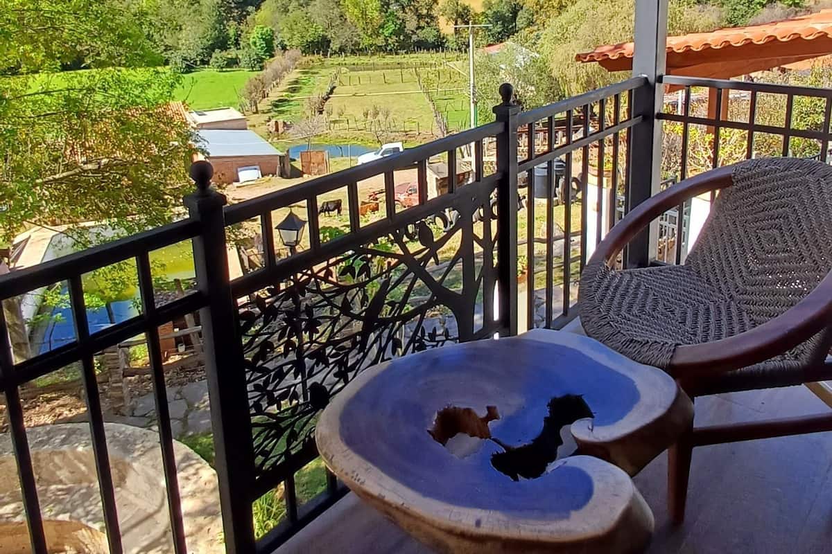 Un balcón con sillas de mimbre y una mesa, con vistas a un paisaje rural con campos y colinas.