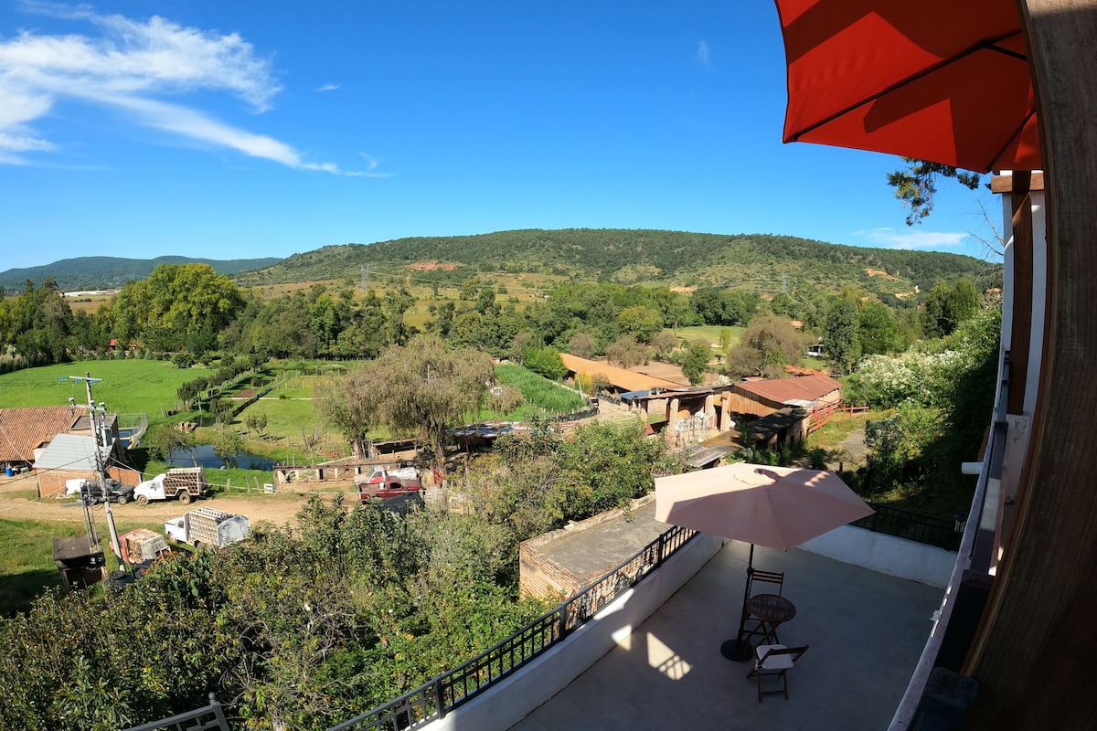 Un balcón con una sombrilla roja, con vista a un valle verde con casas y árboles.