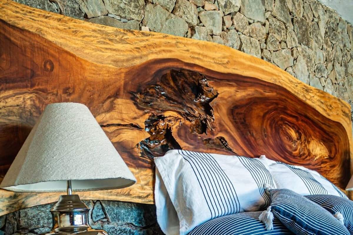 Una cama con ropa de cama a rayas, una lámpara sobre una mesita de noche de madera y un muro de piedra al fondo.