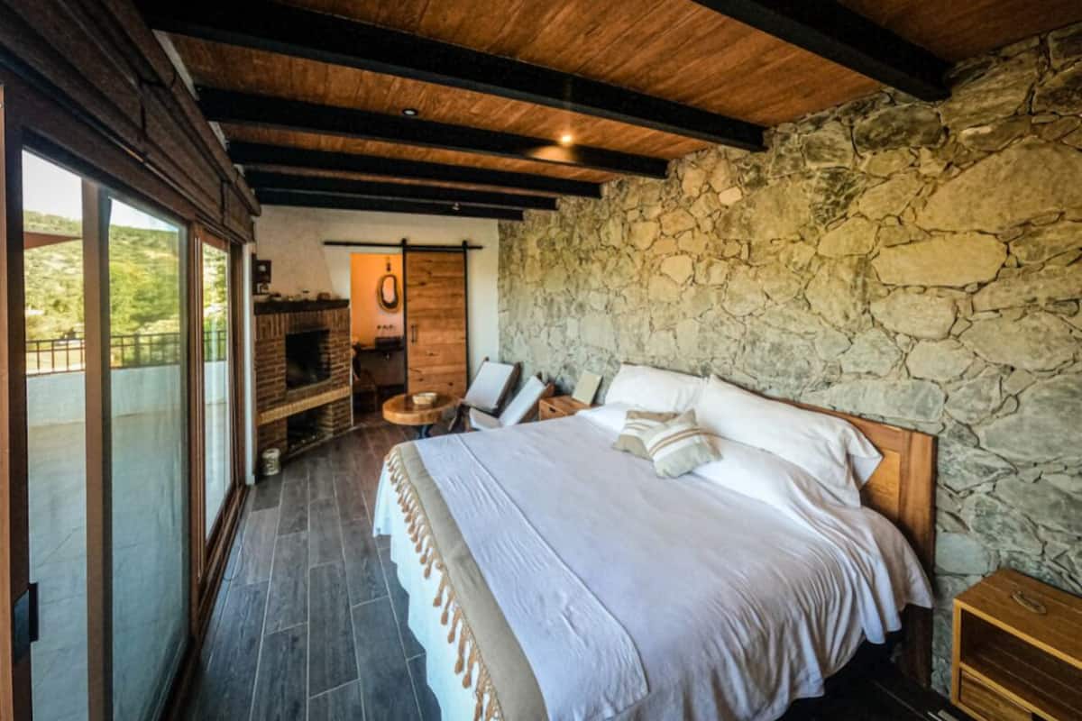 Un dormitorio con pared de piedra, cama con ropa de cama blanca, techo de madera y chimenea.