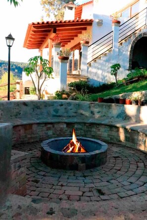 A fire pit in a circular stone design, surrounded by a paved area with a stone wall and a building with a red-tiled roof in the background. - La Toscanía Tapalpa Resort & Agroturismo (Tapalpa)
