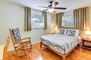 Un dormitorio con una cama de madera, una mecedora, un ventilador de techo y dos ventanas con cortinas.