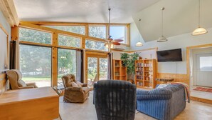 A spacious living room with large windows, a ceiling fan, a flat-screen TV, and a bookshelf. - Cozy Idaho Retreat w/ Grill & Fire Pit! (Caldwell)