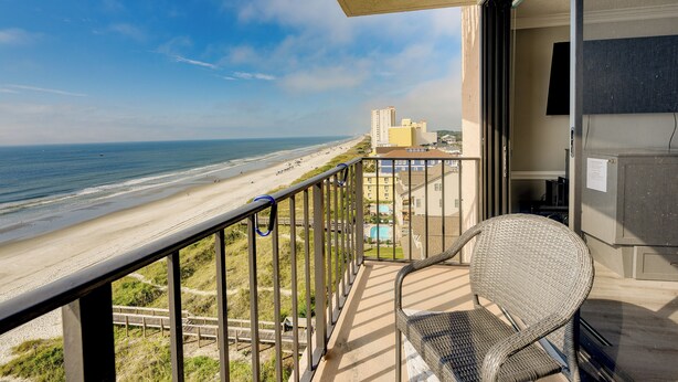 A balcony with a wicker chair, overlooking a beach with a clear sky. - Pool Access: 8th-floor Condo on North Myrtle Beach (North Myrtle Beach)