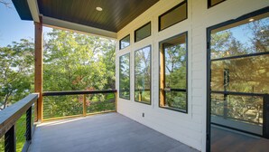 A covered outdoor balcony with large windows, wooden railings, and a view of trees. - Modern Kingsland Home w/ River Access & Kayaks! (Kingsland)