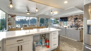 A modern kitchen with a large island, white cabinetry, and pendant lighting. There are wicker baskets on the island, and a fire extinguisher is visible. - Waterfront Allyn Retreat w/ Guest House & Beach! (Allyn)