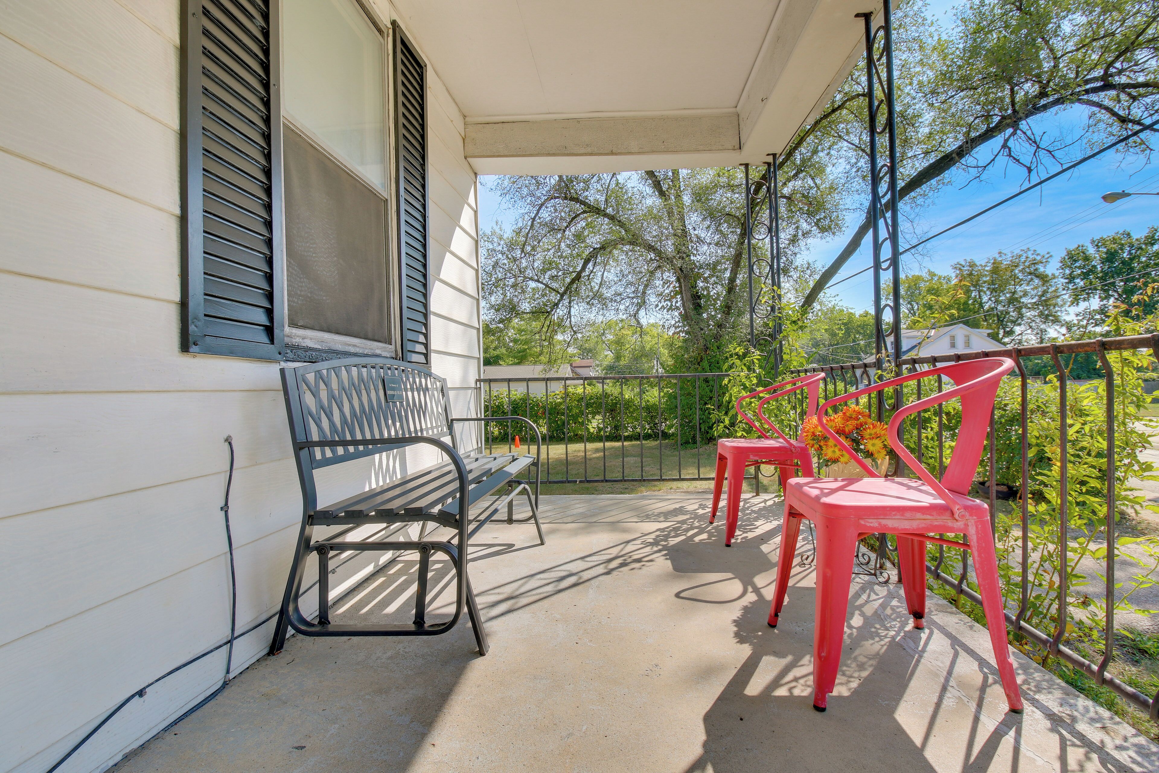 A covered porch with a bench and two chairs, a railing, and a view of trees and a house in the background.