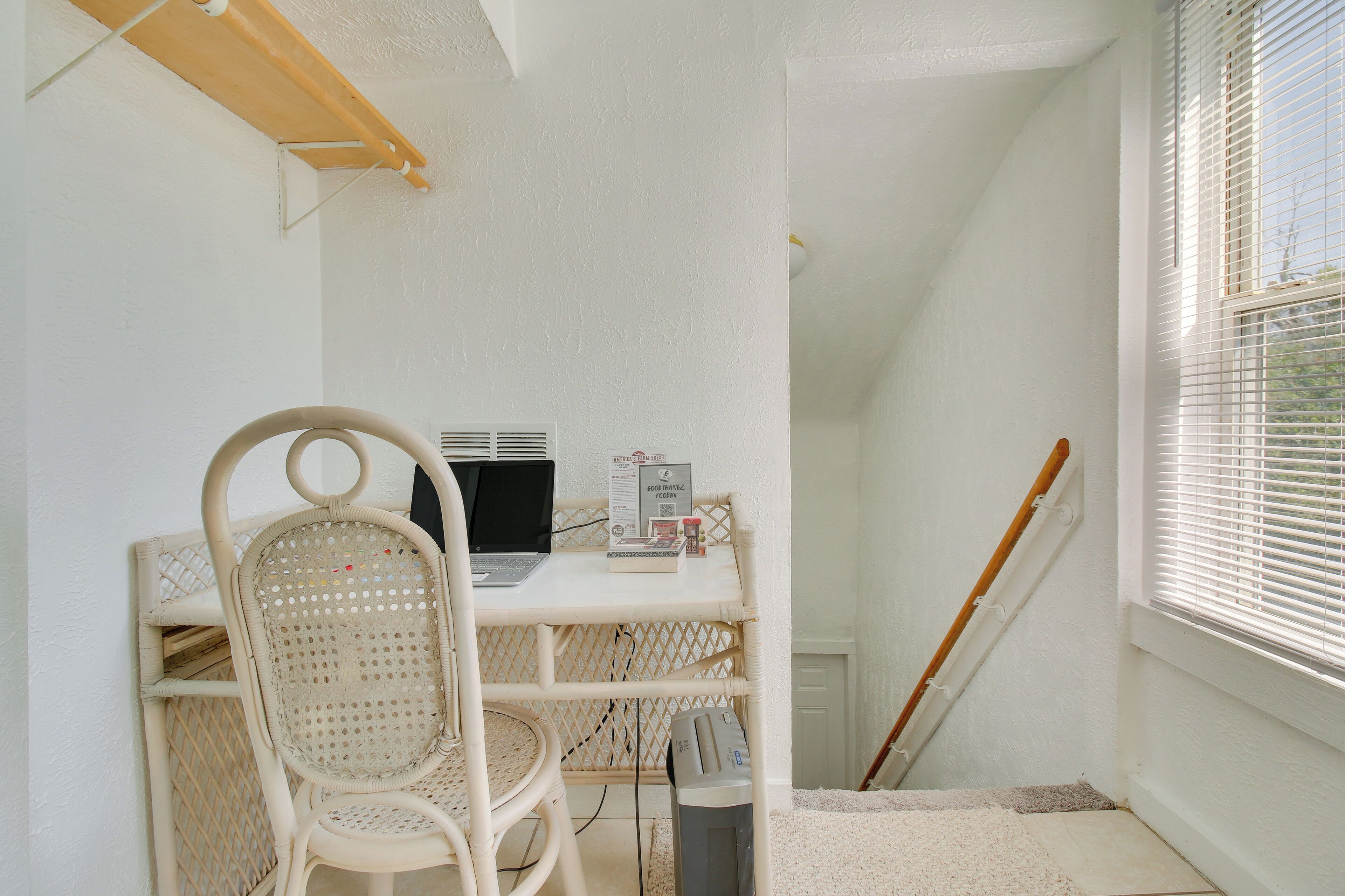 A white-walled room with a white desk, a wicker chair, a laptop, and a broom leaning against the wall.