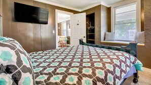 A bedroom with a patterned bedspread, a flat-screen TV mounted on the wall, a window with blinds, and a ceiling fan.