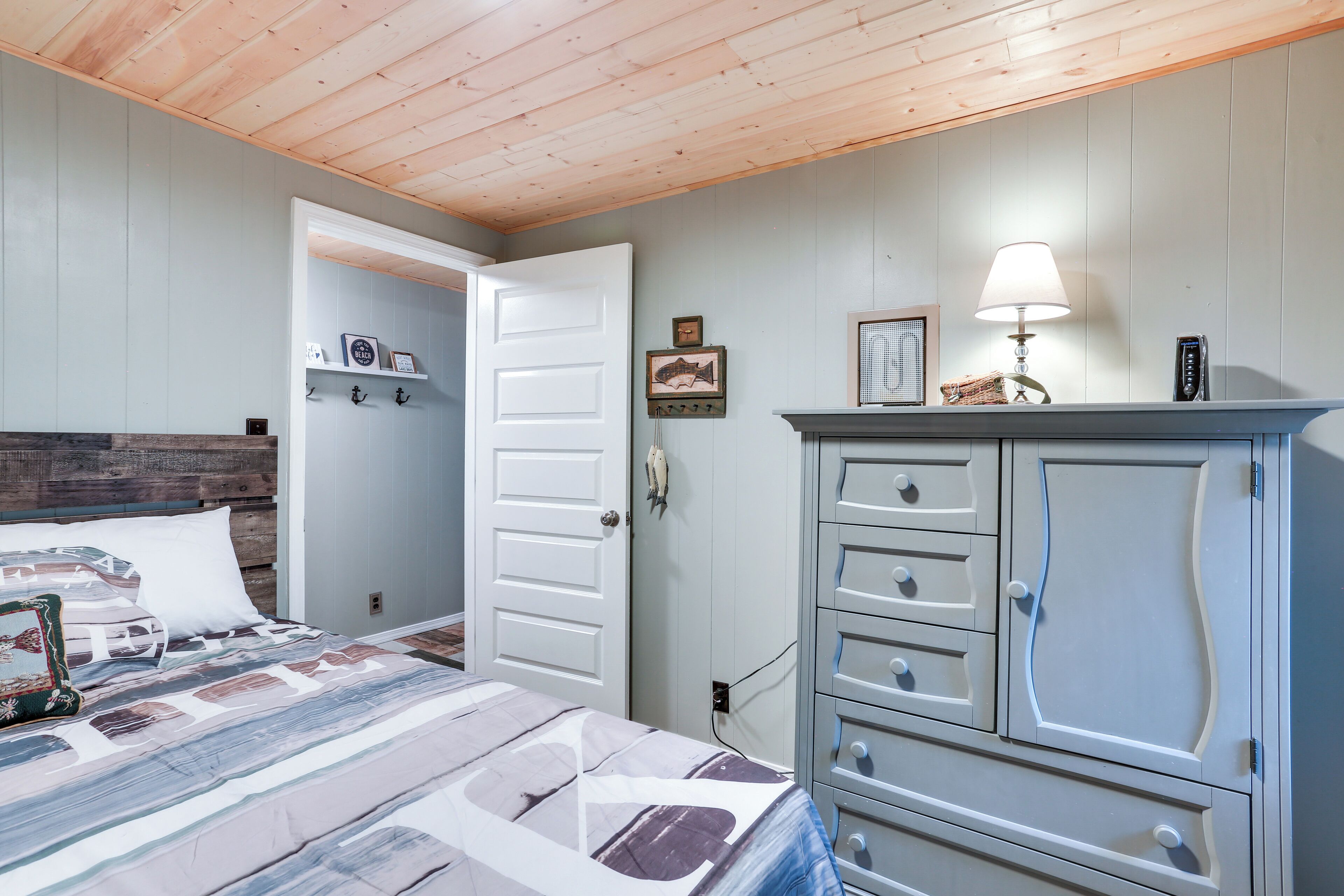 Un dormitorio con cabecera de madera, cama con colcha estampada, cómoda con cajones, lámpara y puerta que da a otra habitación.