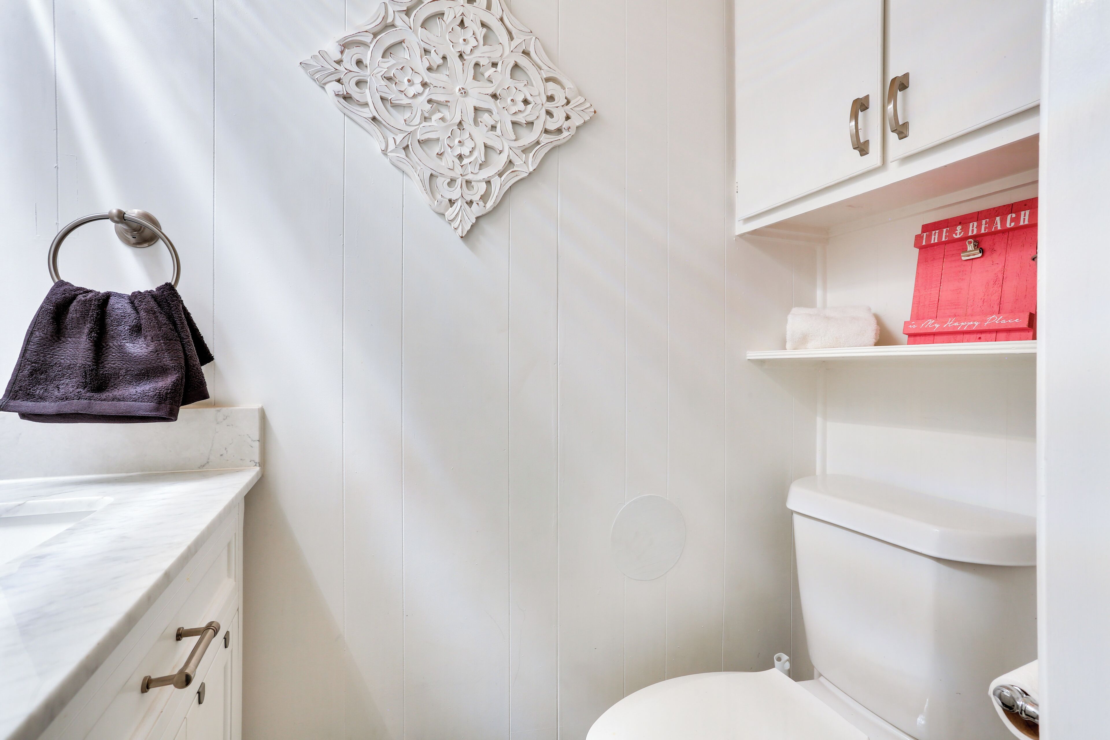 Une salle de bain avec un meuble-lavabo blanc, un porte-serviettes, un élément décoratif mural, des toilettes et une boîte de rangement rouge portant l’inscription « The Beach ».