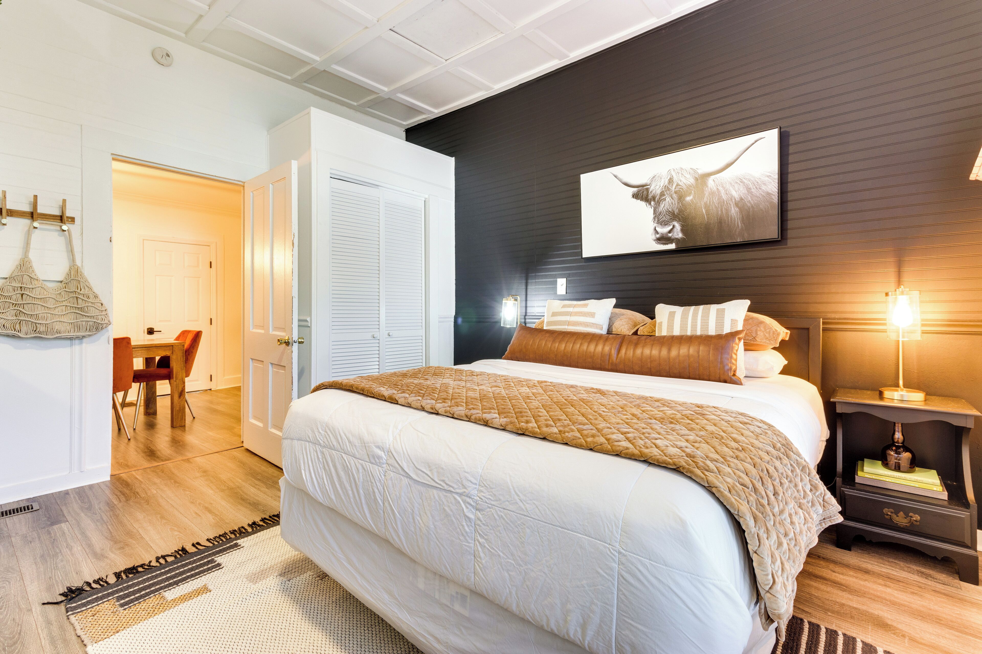 A bedroom with a large bed, a bedside table with a lamp, a wall-mounted picture, and a door leading to another room.