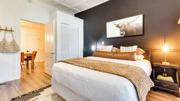 A bedroom with a large bed, a bedside table with a lamp, a wall-mounted picture, and a door leading to another room.