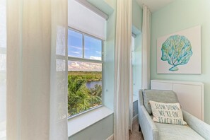 Una habitación con un ventanal grande que da a un paisaje natural, una cama con un almohadón que dice "LOW DOWN WE'RE ON BEACH TIME", y una obra de arte colgada en la pared con forma de hoja.