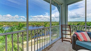 A balcony with a view of a river and greenery, a wicker sofa with a striped pillow, and a metal railing. - Serenity on the Sound, steps to pool/grills (Bradenton)