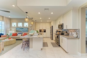 A modern kitchen with white cabinets, granite countertops, and stainless steel appliances. There is a dining area with a round table and chairs, and a living room with a sofa and a coffee table. - Serenity on the Sound, steps to pool/grills (Bradenton)