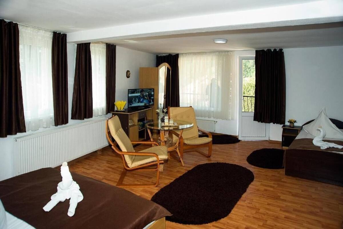 Quarto de hotel com cama, duas poltronas, uma mesinha, televisão e janela com cortinas.