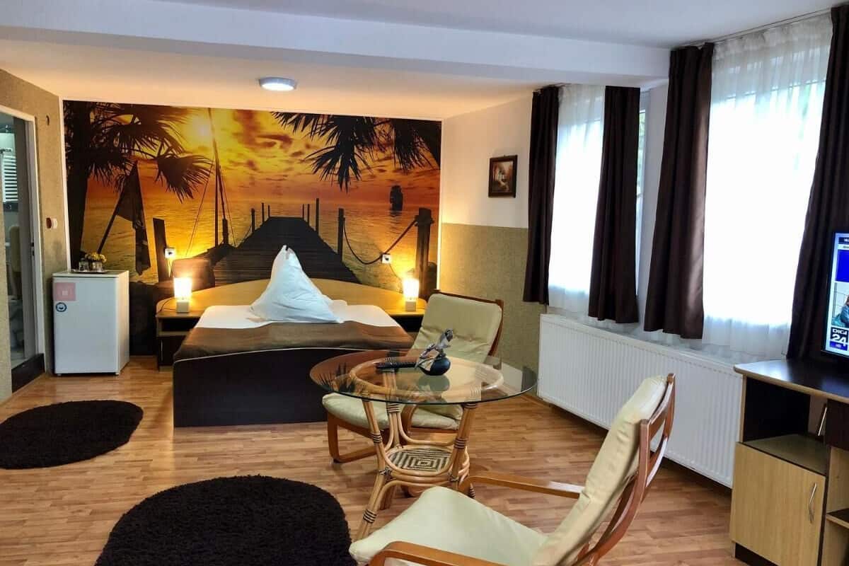 Quarto de hotel com cama, mesa de vidro, duas cadeiras, uma televisão pequena e um mural com um pôr do sol.
