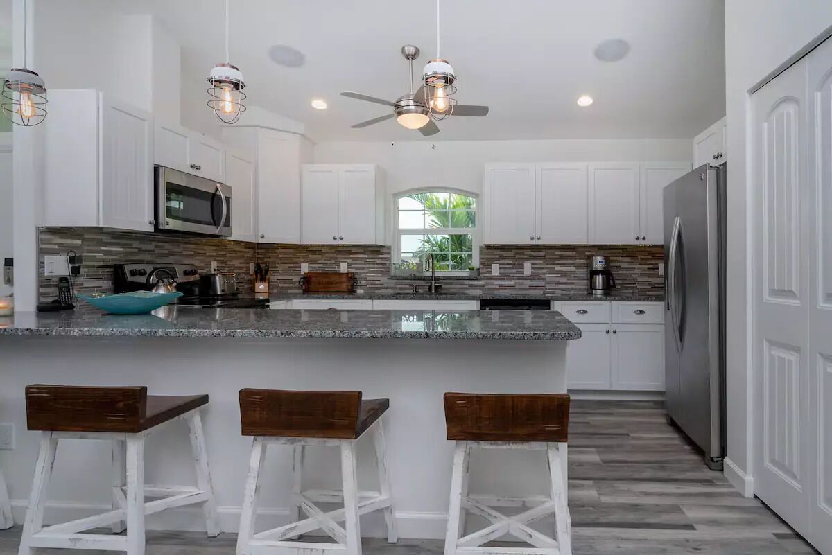 A modern kitchen with white cabinets, a granite countertop, a built-in microwave, a stainless steel refrigerator, and a ceiling fan.