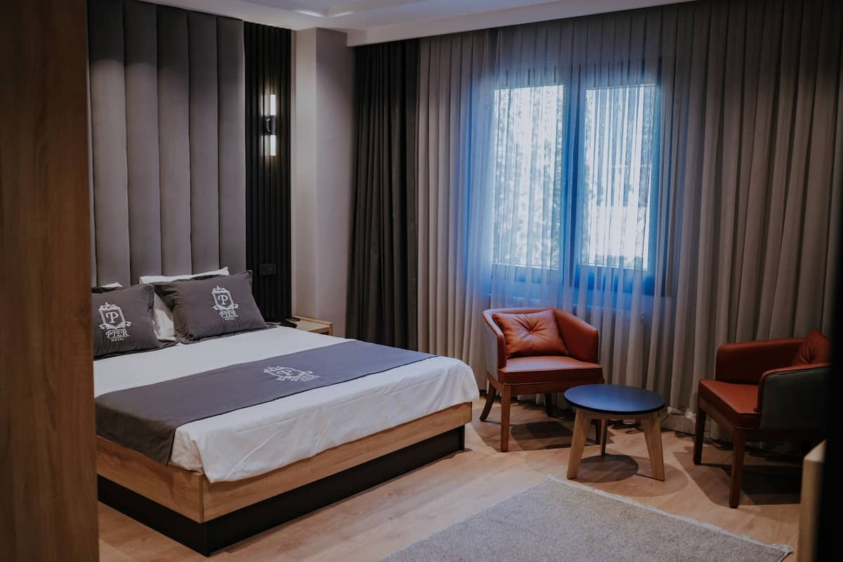 A modern hotel room with a large bed, a small round table, two armchairs, and a window with curtains.
