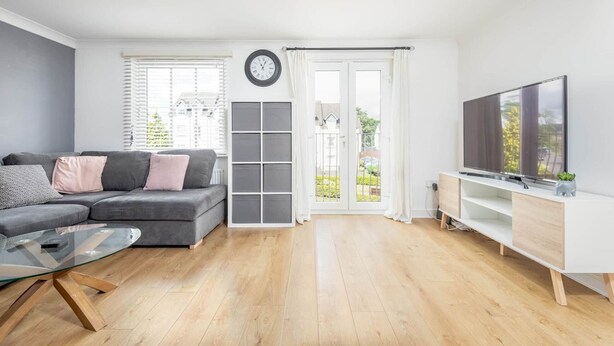 A modern living room with a grey sofa, a glass coffee table, a TV on a white stand, and a balcony with a view. - Chambers Place (No 10) (St Andrews)