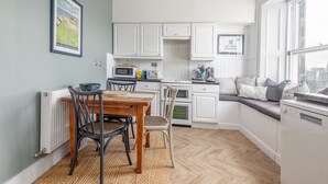 Une cuisine compacte avec des meubles blancs, une table à manger avec des chaises, un banc intégré avec des coussins et une vue par la fenêtre.