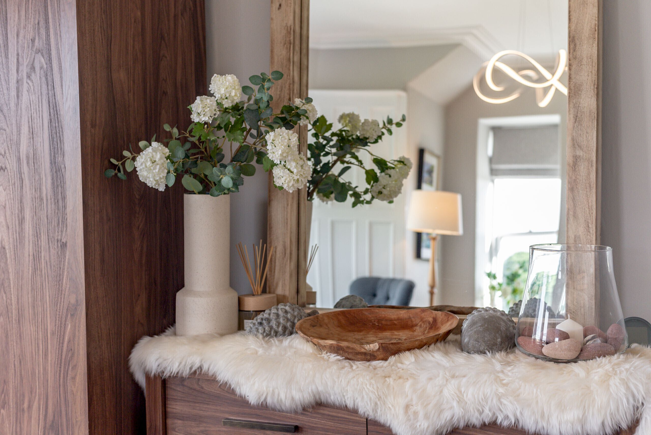 A wooden dresser with a white furry rug, a vase with white flowers, a wooden bowl, and a glass candle holder.