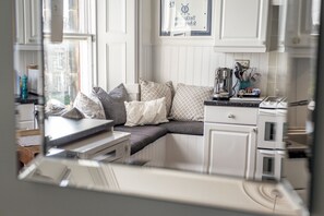 Une cuisine avec des meubles blancs, un banc intégré avec des coussins, une machine à café et une vue par la fenêtre.