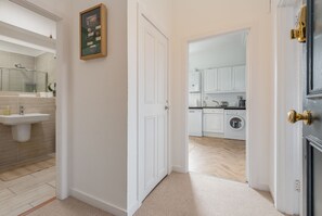 A modern bathroom with a sink, mirror, and shower. A hallway leads to a kitchen with cabinets and a washing machine.