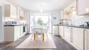 A modern kitchen with white cabinetry, a dining area with a white table and chairs, and a view through an open door to the outdoors.