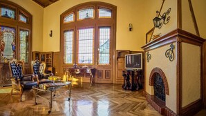 Un vaste salon doté d’un parquet en bois disposé en point de Hongrie, d’une cheminée et de grandes fenêtres ornées de vitraux.