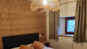 A bedroom with a wooden headboard, a bed with white linens and two brown pillows, a grey throw blanket, a woven light fixture, a window with curtains, and a radiator. - Ferme Ancienne 1876 Rénovée, Appartement Neuf Tout Confort 8 Personnes (Le Grand-Bornand)