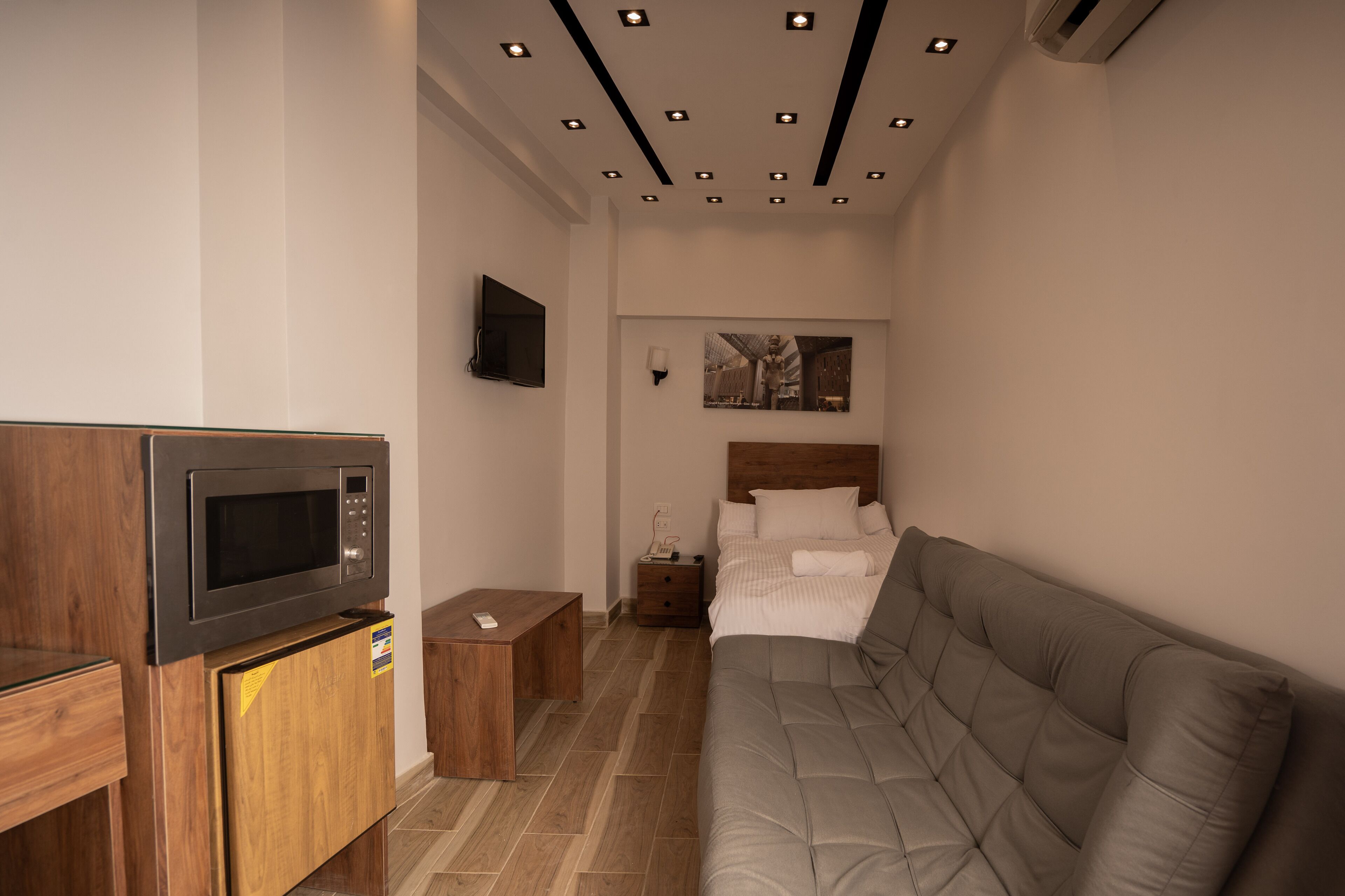 Una camera d'albergo moderna con forno a microonde integrato, un armadietto in legno, un comodino, un piccolo tavolino da notte, un letto con testiera e un soffitto con faretti.
