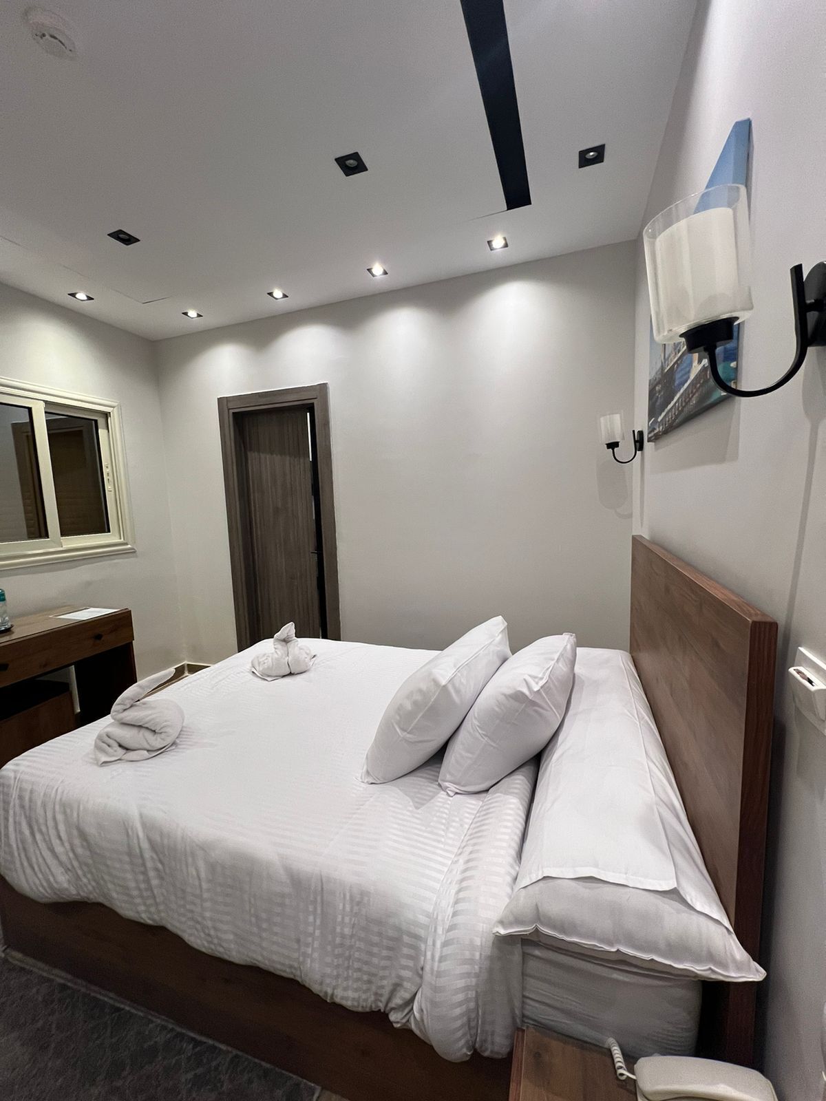 Un letto ben rifatto con lenzuola e cuscini bianchi, una testiera in legno, un comodino con una lampada e uno specchio sulla parete.