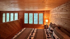 Una camera accogliente con pareti e soffitto in legno, un letto grande con copripiumino decorato, un comodino con lampada e ampie finestre che offrono vista sugli alberi.