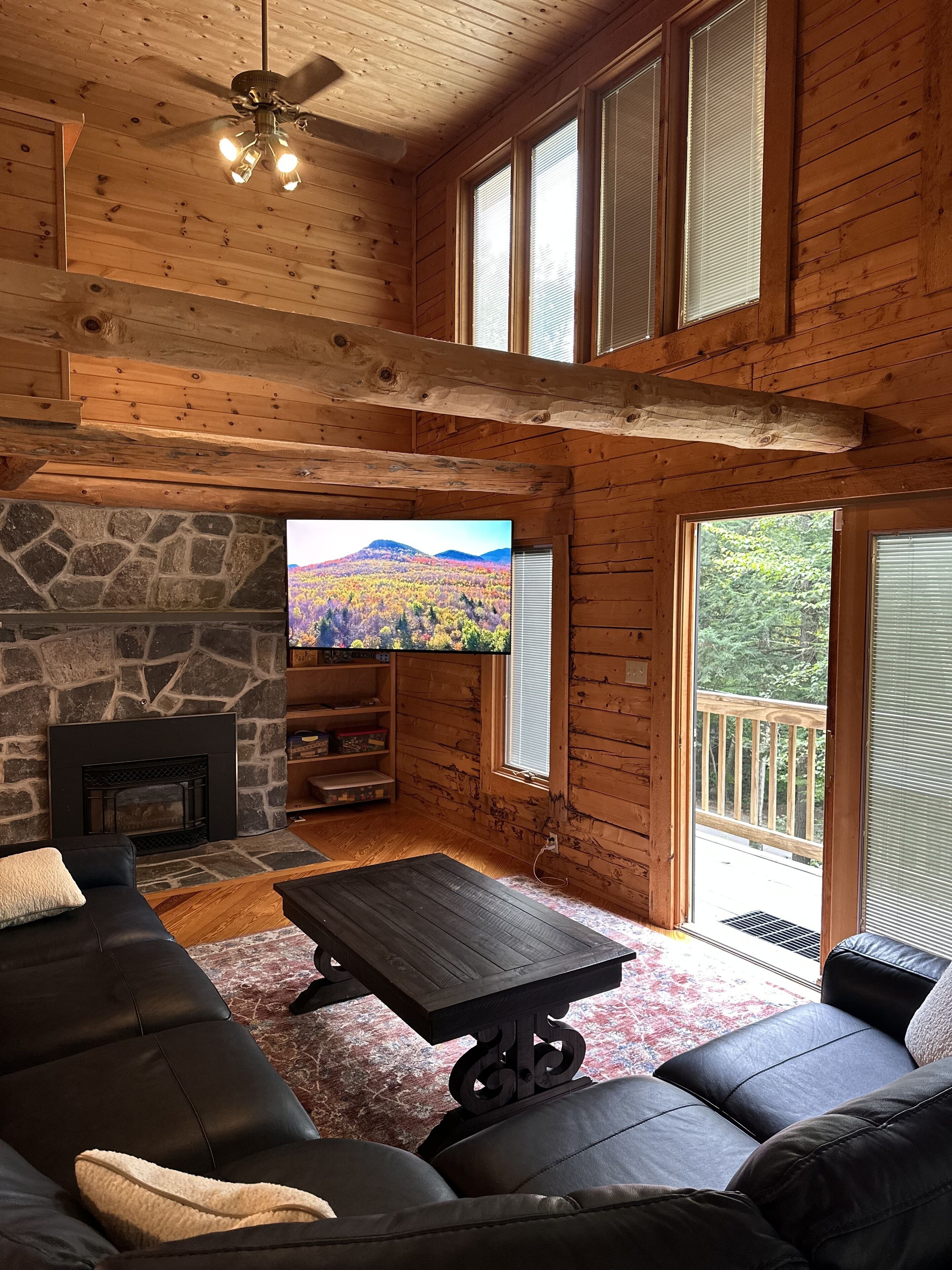 Interior acolhedor de uma cabana com lareira de pedra, um sofá de couro preto, uma mesa de centro de madeira e uma televisão exibindo uma paisagem cênica.