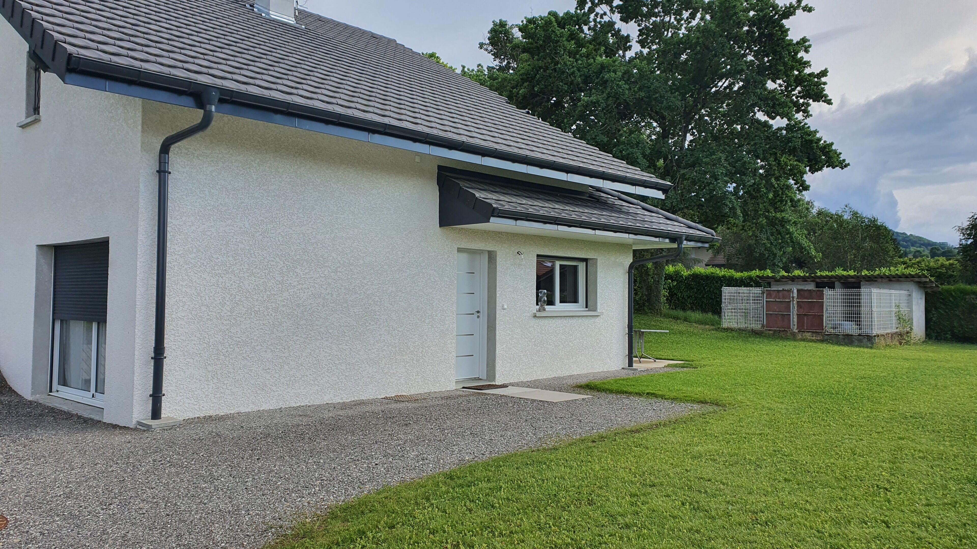 Ein eingeschossiges Haus mit weißer Fassade, einem grauen Dach und einer Auffahrt mit Kies zum Eingang.
