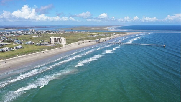 Une vue aérienne d’une zone côtière avec une plage, une jetée s’avançant dans l’eau et un ensemble de bâtiments situés près du rivage.