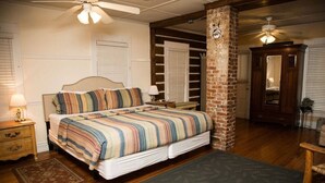 A bedroom with a large bed, wooden flooring, a brick column, and a ceiling fan.