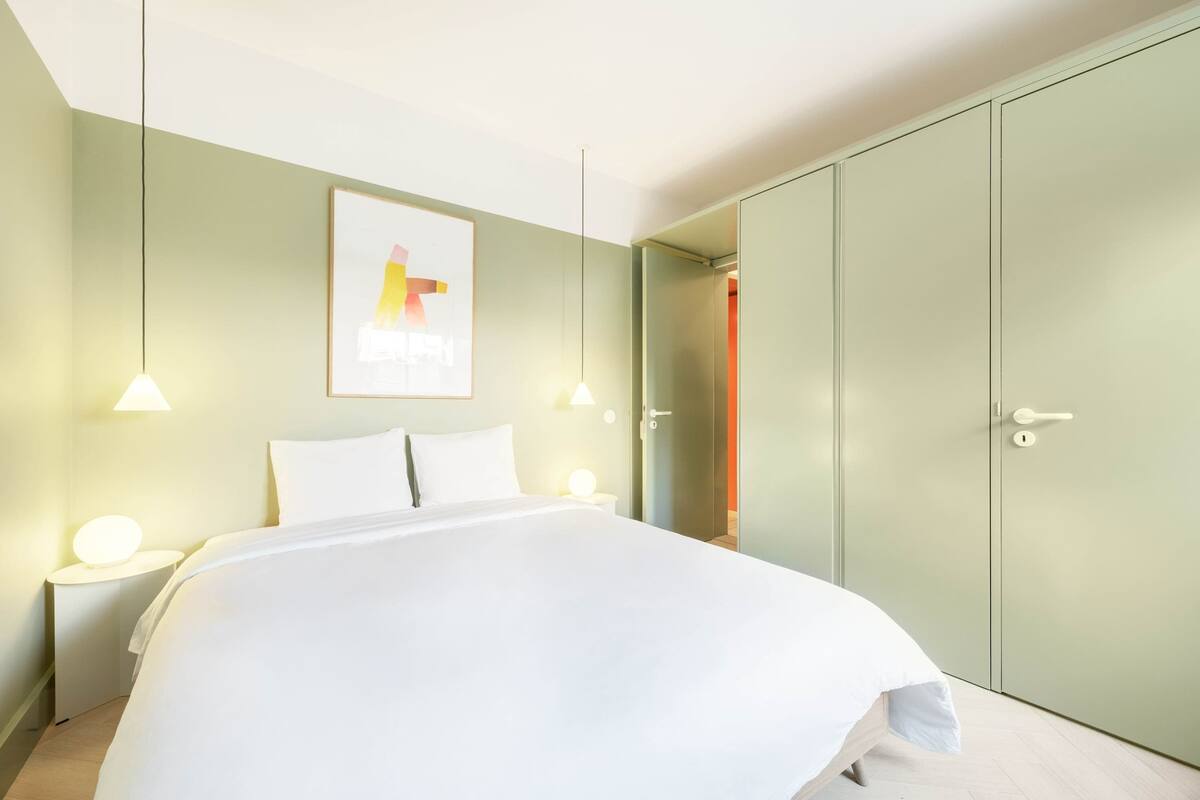 Ein modernes Schlafzimmer mit einem großen Bett, Nachttischlampen, einem Kleiderschrank und einem gerahmten Kunstwerk an der Wand.