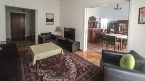 A living room with a sofa, a TV, a table with a tablecloth, a patterned rug, and a chandelier. There is a door leading to another room. - 3-Generations Athenian home at the foot of the Acropolis hill in Athen"s center (Athina)