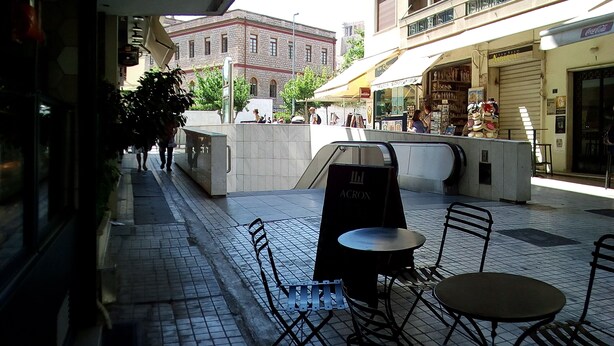 An outdoor seating area with tables and chairs, a sign for 'Acron', and a view of a street with buildings and shops. - 3-Generations Athenian home at the foot of the Acropolis hill in Athen"s center (Athina)