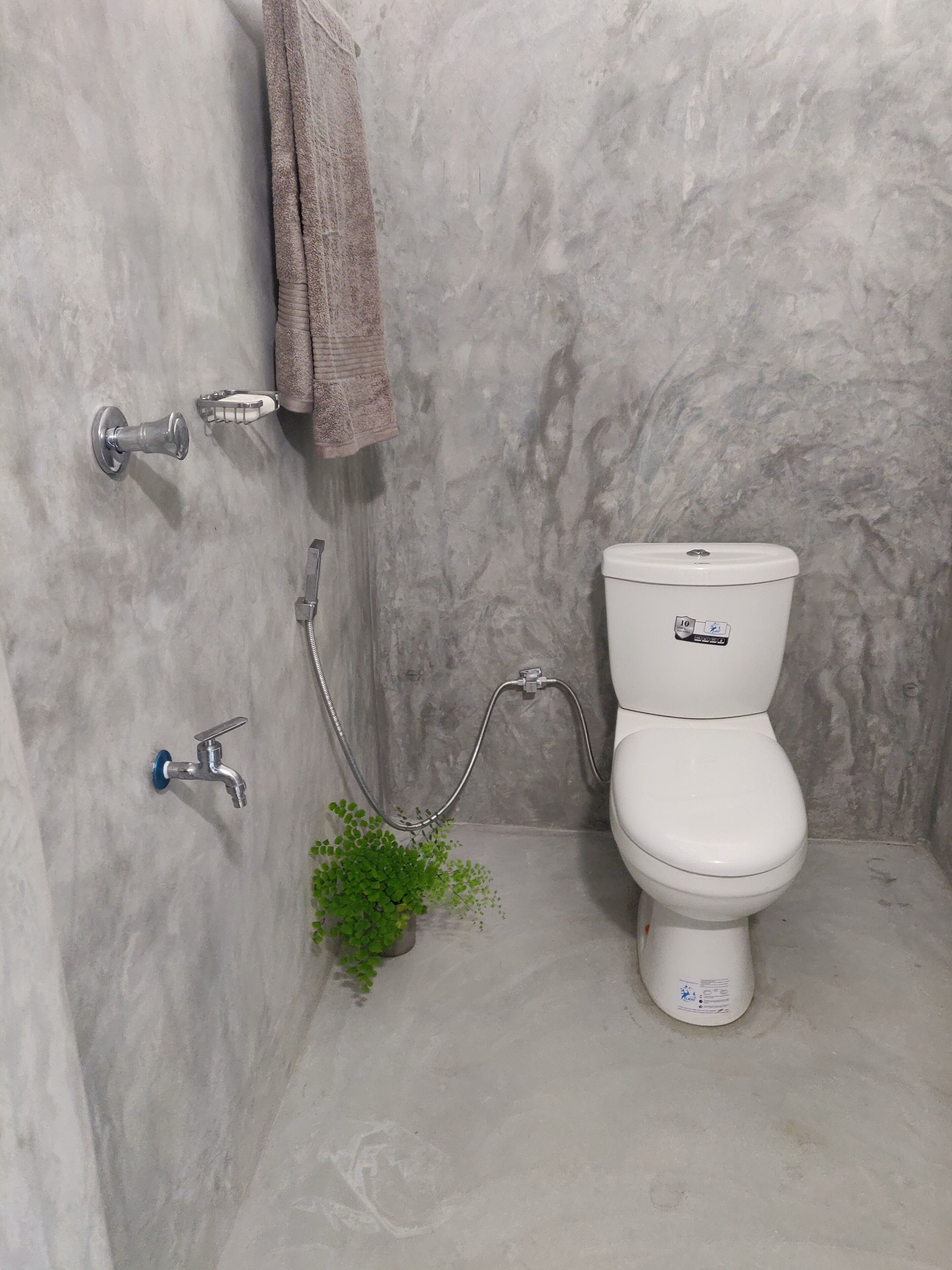 Un baño con inodoro blanco, ducha con cabezal de ducha de mano y una toalla colgada en un perchero.