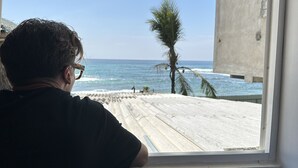 A person looking out of a window at a beach with a palm tree and a concrete building in the background. - Dipndoze (Ahangama)