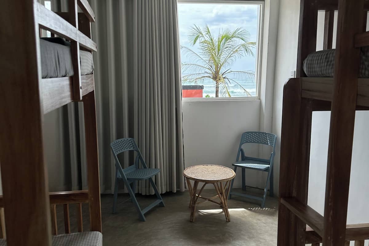 A room with bunk beds, two chairs, a small round table, and a window with a view of the sea and palm tree.